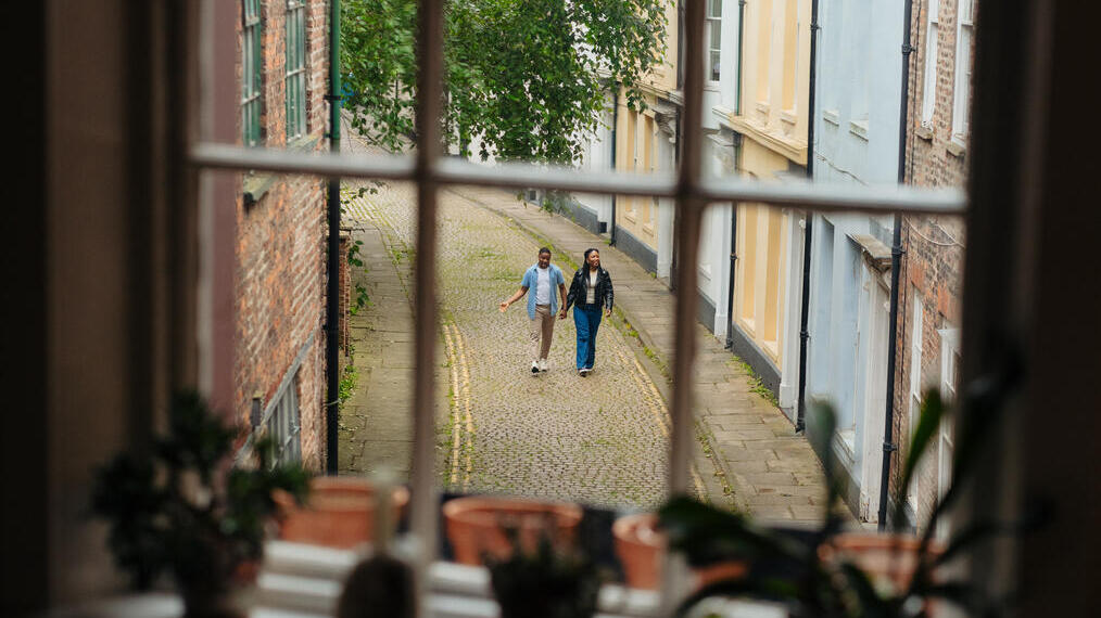 View through a window onto a cobblestone street with two people walking, surrounded by historic brick and pastel buildings.