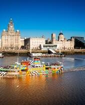 Colorful ferry on the River Mersey with Liverpool waterfront, historic buildings, and modern architecture under a clear blue sky.