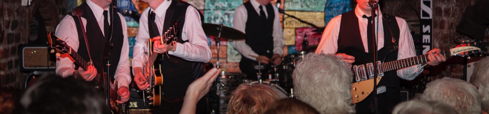A band dressed in matching black vests and ties performs live in a crowded, arched brick venue with a colorful graffiti-style wall in the background.