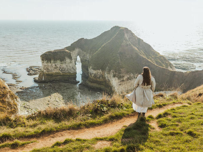 Person in light coat stands on grassy cliff path overlooking dramatic sea stacks and archway at the coastline, with clear ocean views.