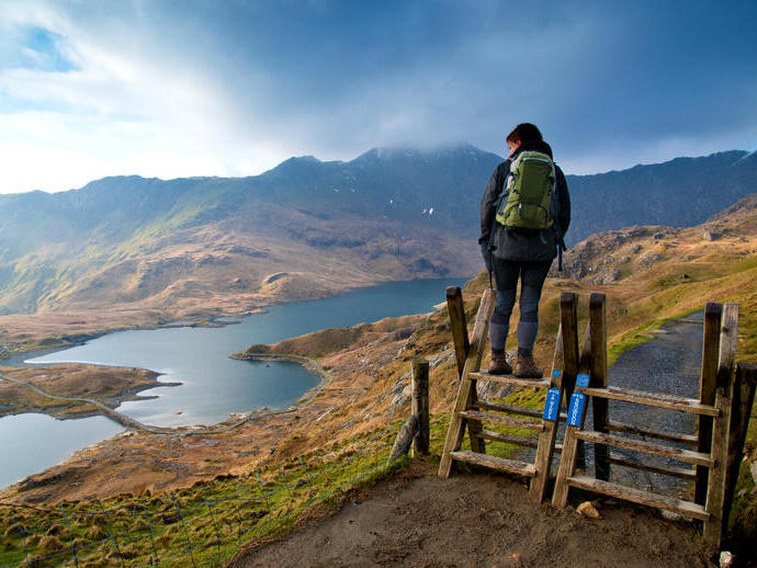 Wanderer auf einem hölzernen Aussichtspunkt mit Blick auf eine weitläufige Bergkette und ein See-Panorama