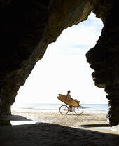 Persona en bicicleta con tabla de surf en la playa, vista desde el interior de una cueva.