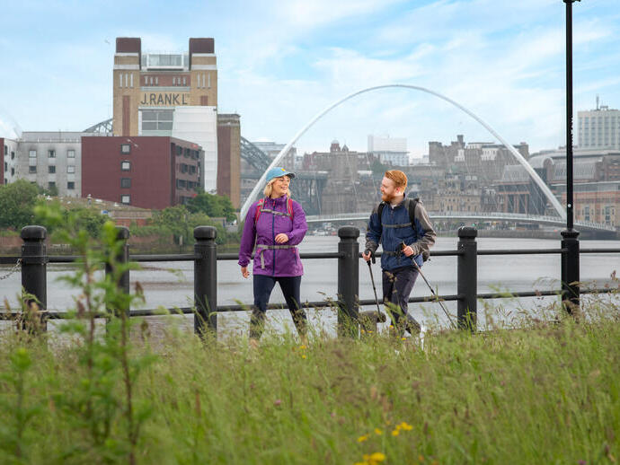 Une femme et un homme marchent le long d'une rivière avec des bâtiments emblématiques et un pont au loin