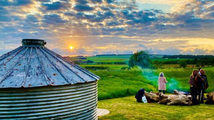 Metal silo, green field at sunset, cloudy sky, and people gathered around a log near a smoking campfire.