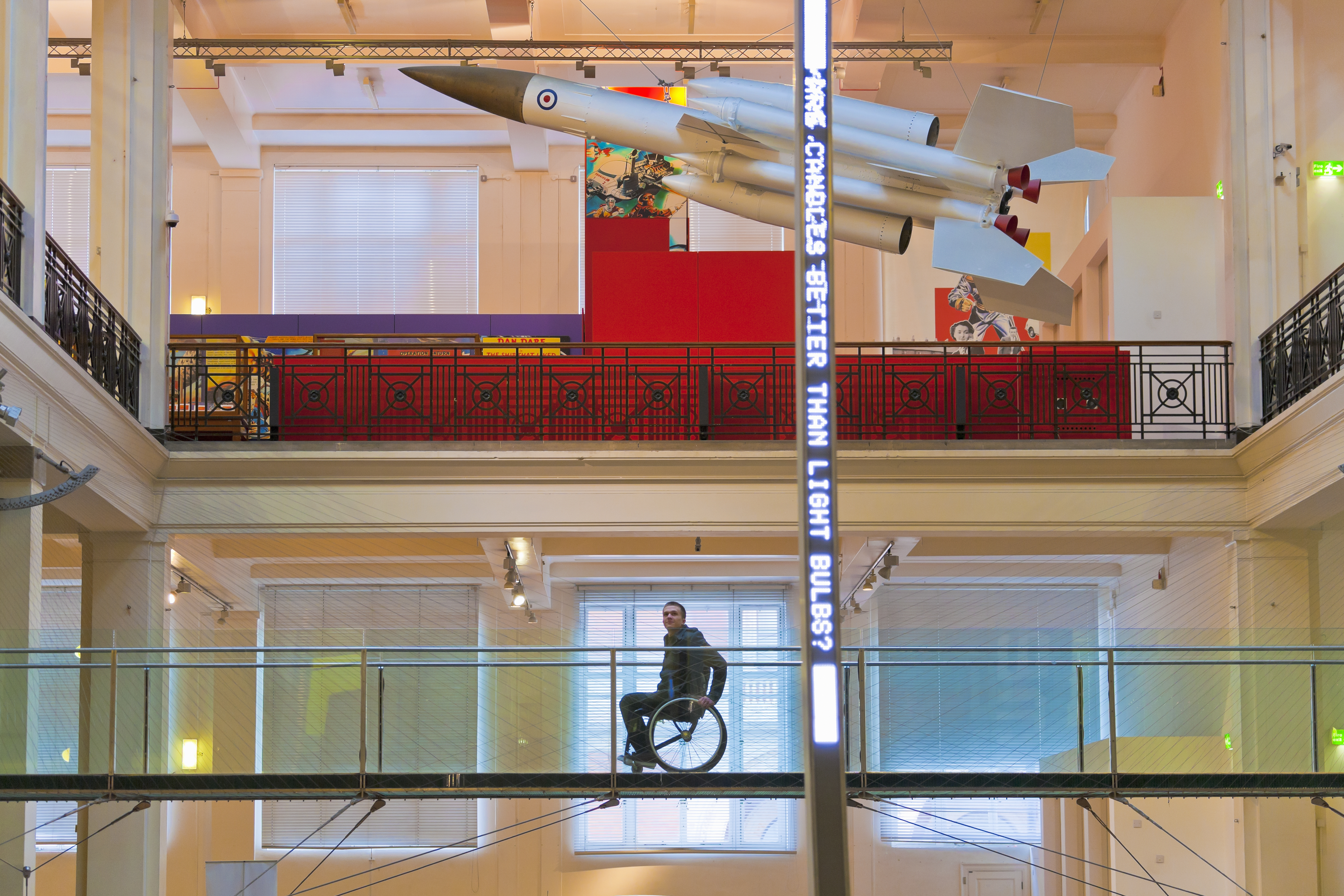 Uomo su una sedia a rotelle su una passerella sopraelevata in un museo che guarda l'esposizione