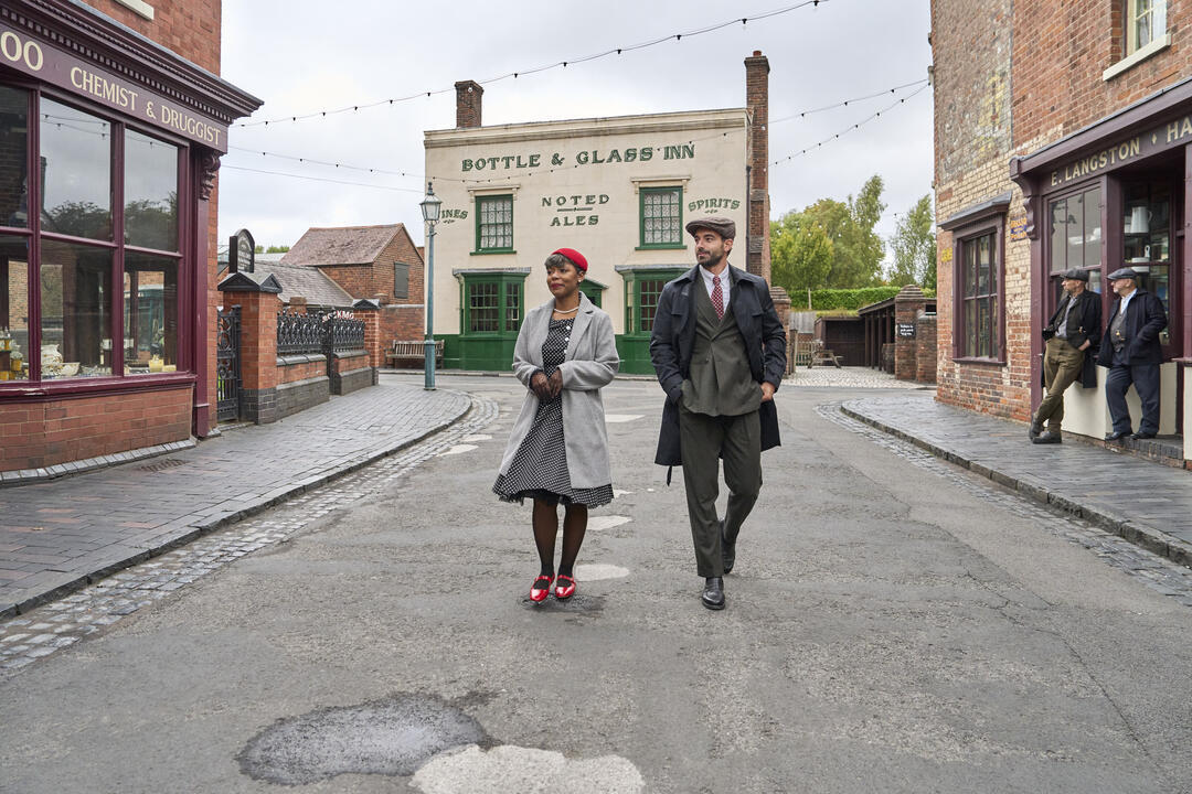 Two people in vintage-style clothing walk down a cobbled street lined with brick shops and an old inn, giving a historical town feel.