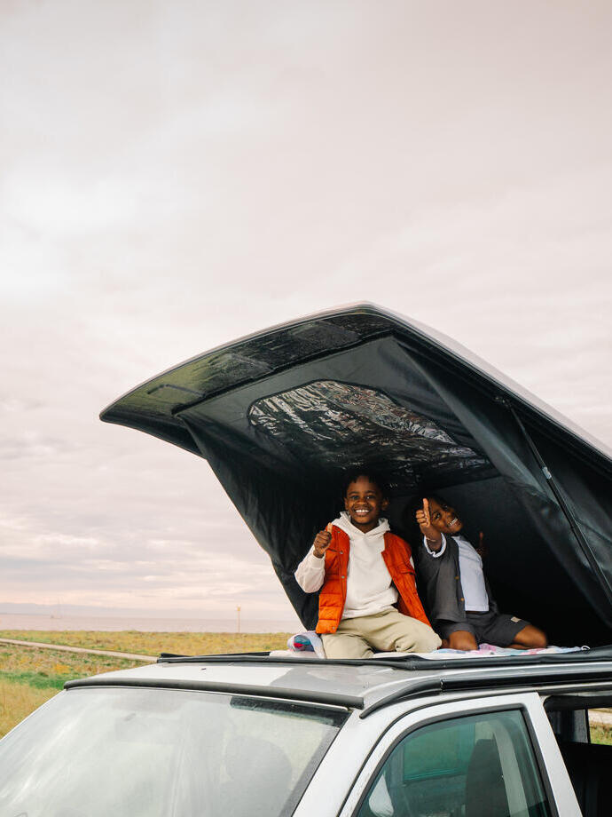 Two children sitting and smiling in a rooftop tent on a car, with grassland and water in the background under a cloudy sky.
