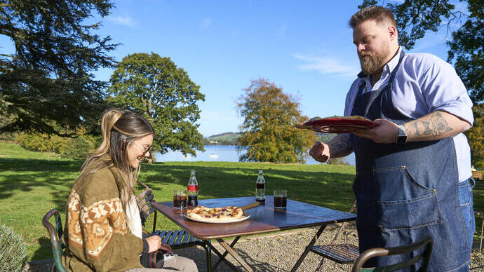Une femme est assise à une table extérieure avec pizza et boissons. Un homme en tablier sert plus de pizza. Paysage vert avec arbres et lac en arrière-plan.