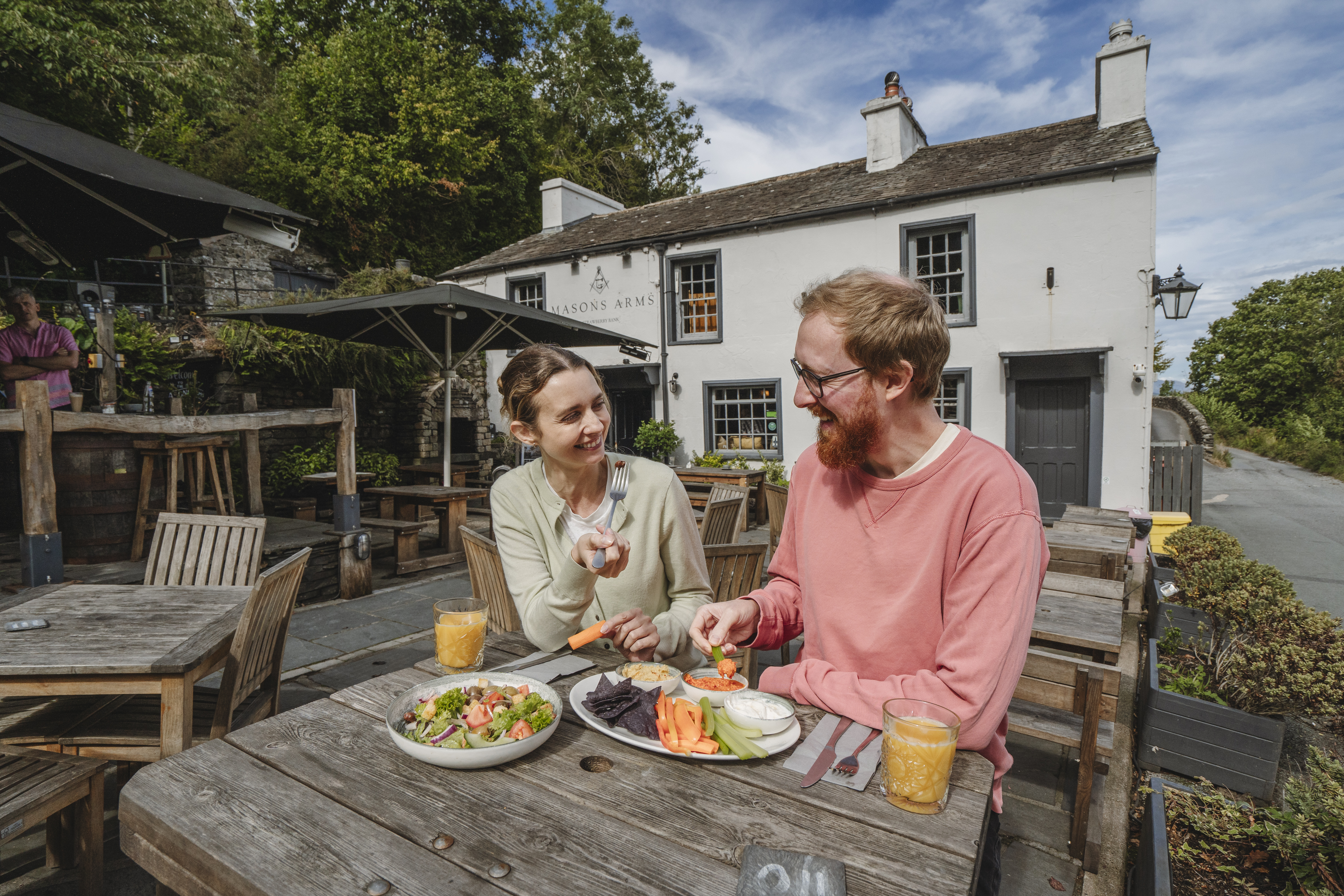Ein Mann und eine Frau sitzen an einem Holztisch im Freien vor einem weißen Gebäude, teilen einen Teller Salat und Gemüsesnacks mit Getränken.