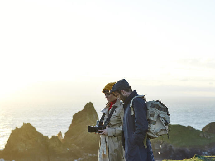 Couple standing on top of the cliffs, the woman holds a camera and the man wears a backpack