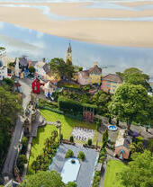 Aerial view of a colorful village with unique architecture, gardens, and a clock tower, set beside a sandy estuary and lush greenery.