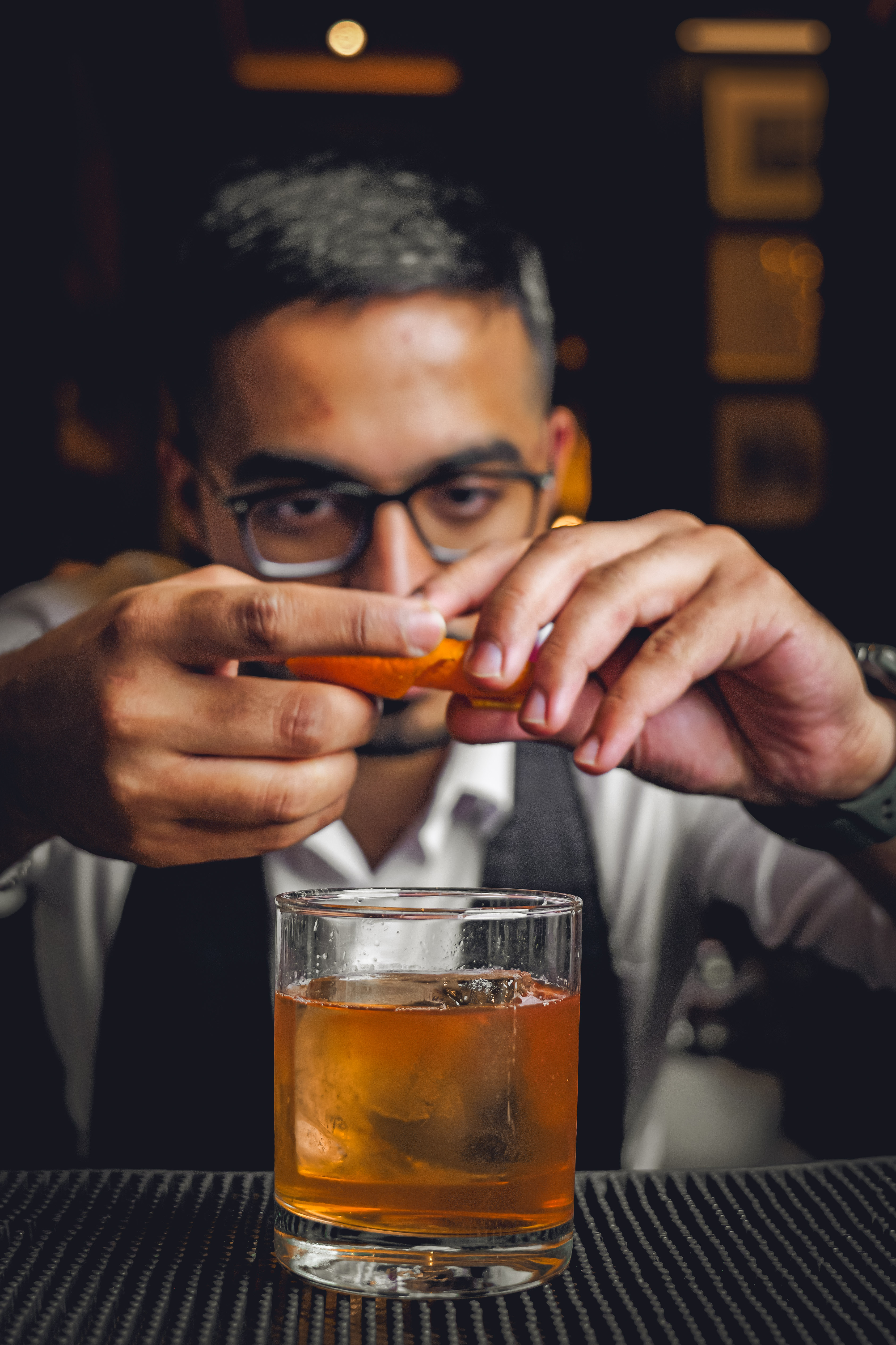 Bartender preparing an orange cocktail garnish above a glass of whiskey or cocktail on a bar counter in a dimly lit setting.