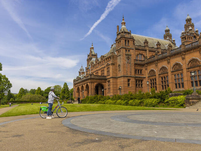 Persona con bicicleta junto a un edificio histórico de ladrillo rojo bajo un cielo azul en un día soleado.