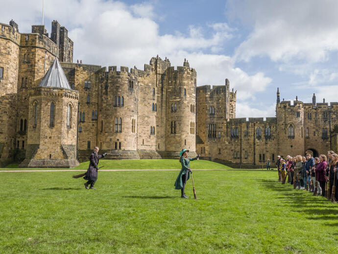 A group of people lined up for a Broomstick Training lesson with the wizarding professors by a large castle.