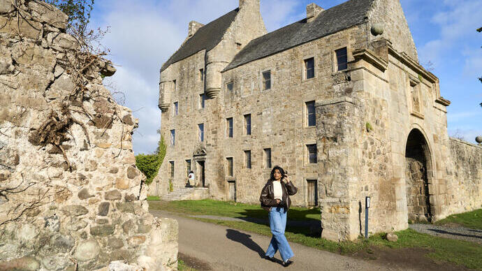 Person walks near a large historic stone building with arched entrance and ruins, under sunny blue sky.