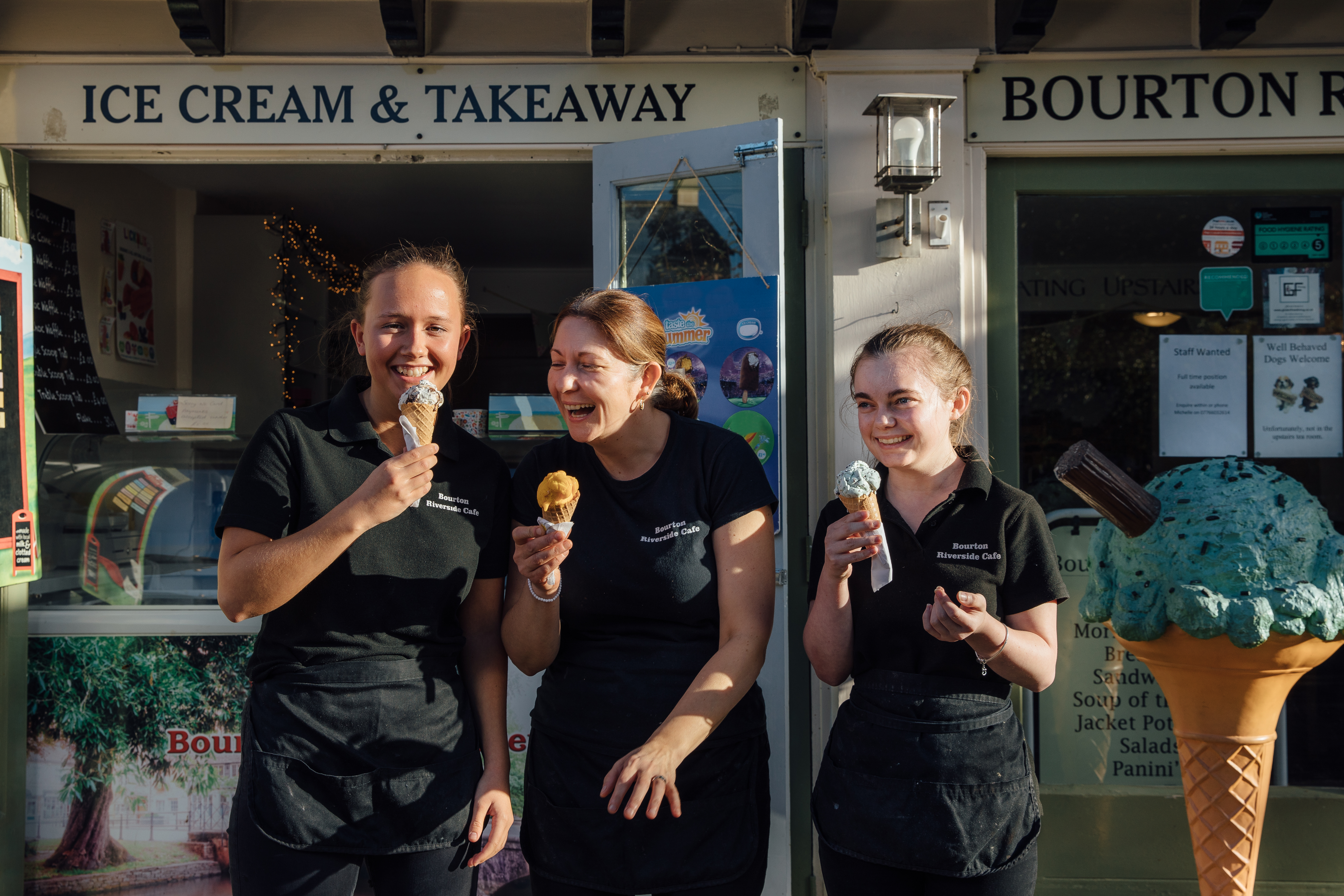 Three smiling woman standing outside ice cream parlour.