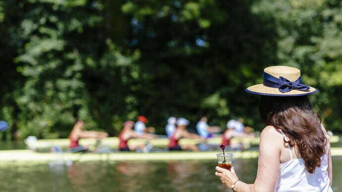 Une femme portant un chapeau, buvant tout en regardant une course de bateaux