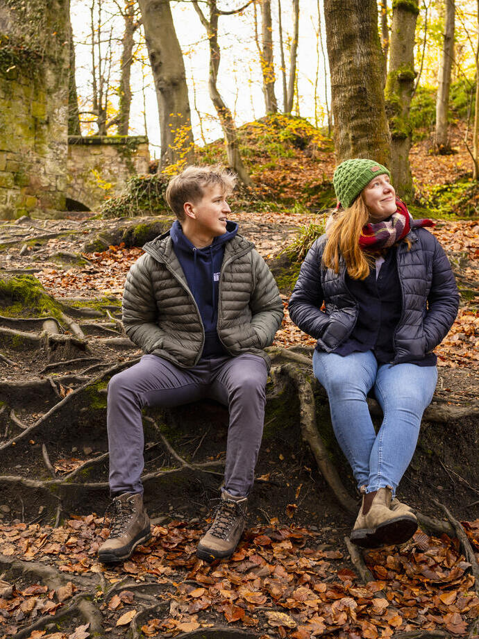 A man and woman in hiking gear smile at each other while sitting on tree roots in a forest.