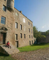 Historic stone building with arched entry gate, green lawn, and cobbled path. A person sits on steps in afternoon sunlight.