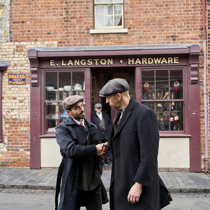 Two men in vintage attire shake hands outside a brick hardware store named E. Langston. Historical street scene with older shopfronts.